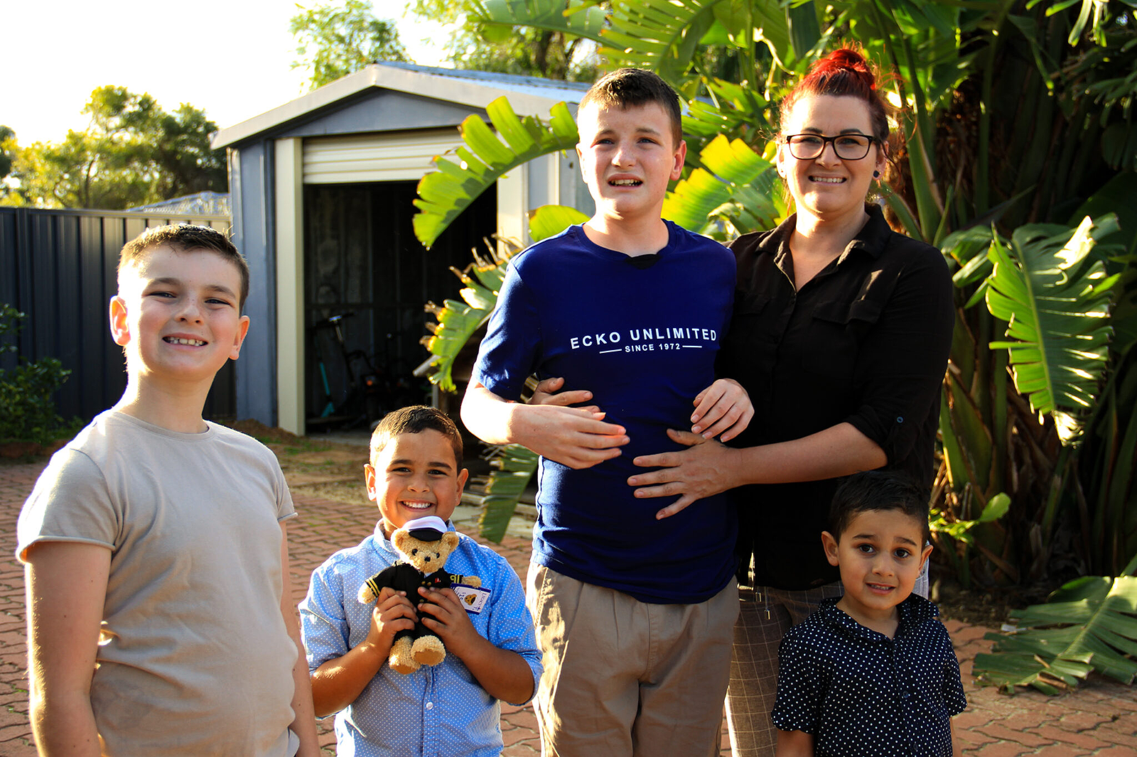 Heidi and her family of four kids stand in a driveway in front of a shed with an open roller door. Tall green plants are in the background.