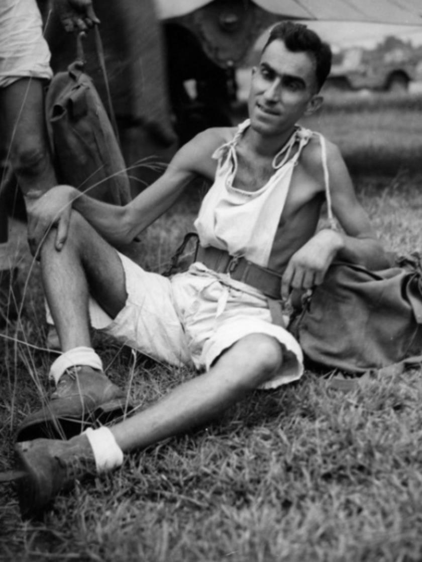 A malnourished man named George Vanselow who is sitting on the ground and leaning back slightly. He is wearing a singlet, shorts, socks and boots.